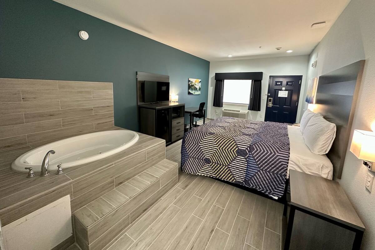 A modern hotel room with a large bathtub, a bed with patterned bedding, a TV, and a desk with a chair.
