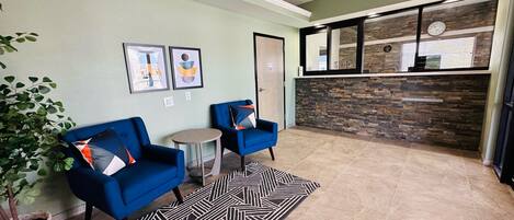 A lobby with two blue armchairs, a small round table, a patterned rug, and a potted plant.