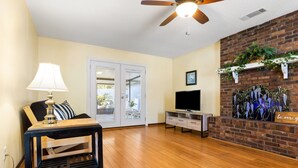 A living room with a wooden floor, a ceiling fan, a TV, a lamp, and a brick wall with a plant shelf. - Enjoy Southern Hospitality in Kingland Georgia, Bring Pets, Plenty Parking (Kingsland)