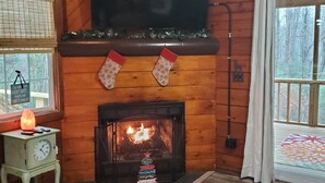 Interno accogliente di una baita con camino, pareti in legno, televisore montato sopra e una finestra con persiane.