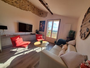 Ein Wohnzimmer mit einer Couch, roten Sesseln, einem kleinen Tisch, einem Fernseher und einem Balkon mit Blick.