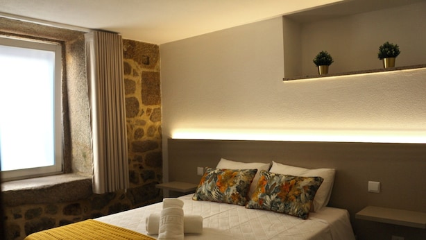 A hotel room with a bed, a window with curtains, a stone wall, a shelf with two small plants, and a bedside table. - Plátanos & River Views I (Ponte de Lima)
