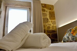 A neatly made bed with a rolled white towel and a colorful pillow, a stone wall, and a window with curtains.