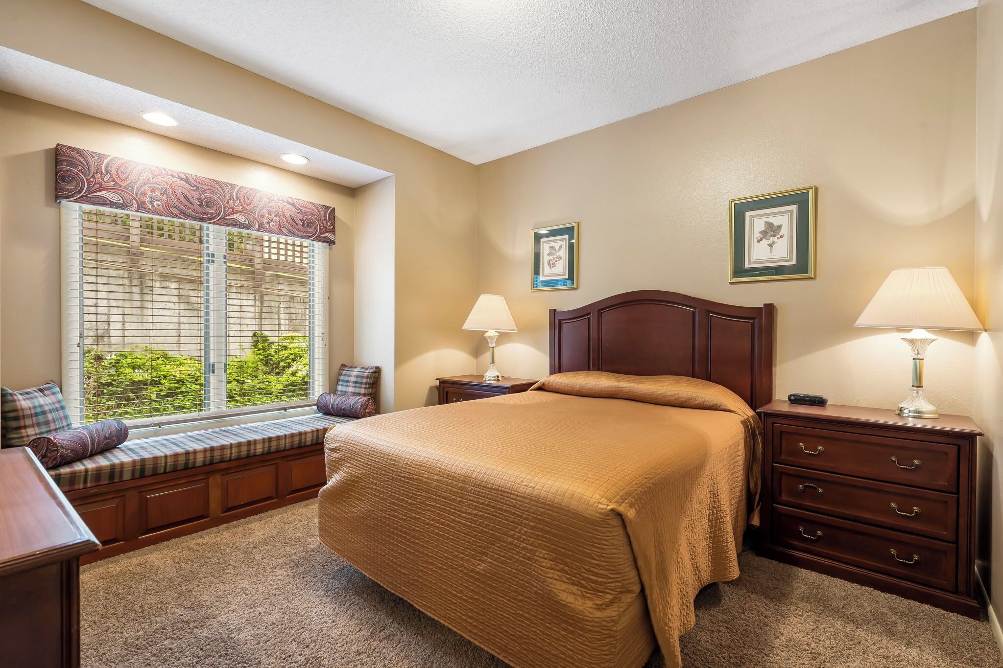 A bedroom with a large bed, a window seat, two bedside tables with lamps, and a dresser.