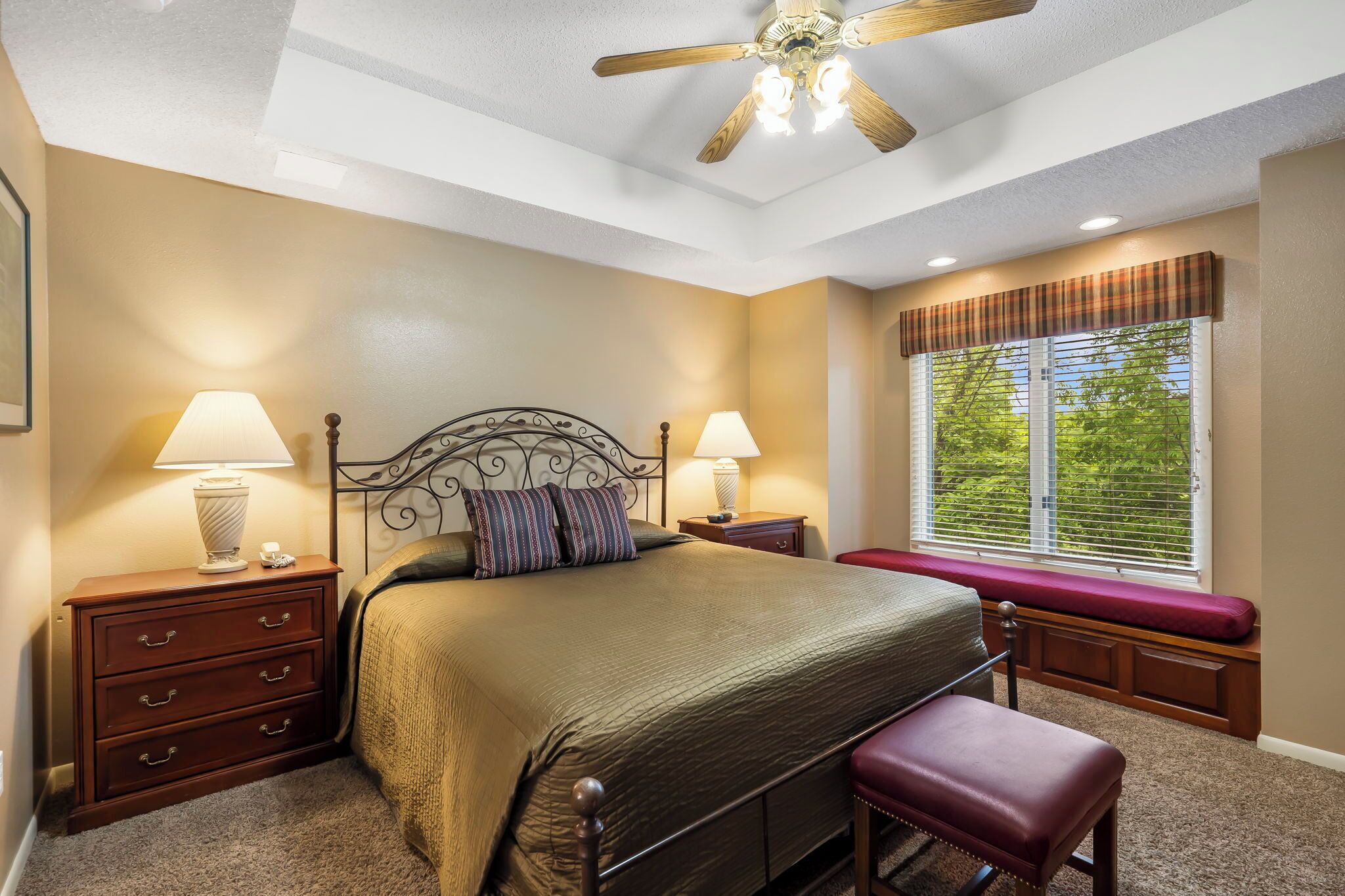 A bedroom with a bed, two bedside tables with lamps, a bench at the window, and a ceiling fan.