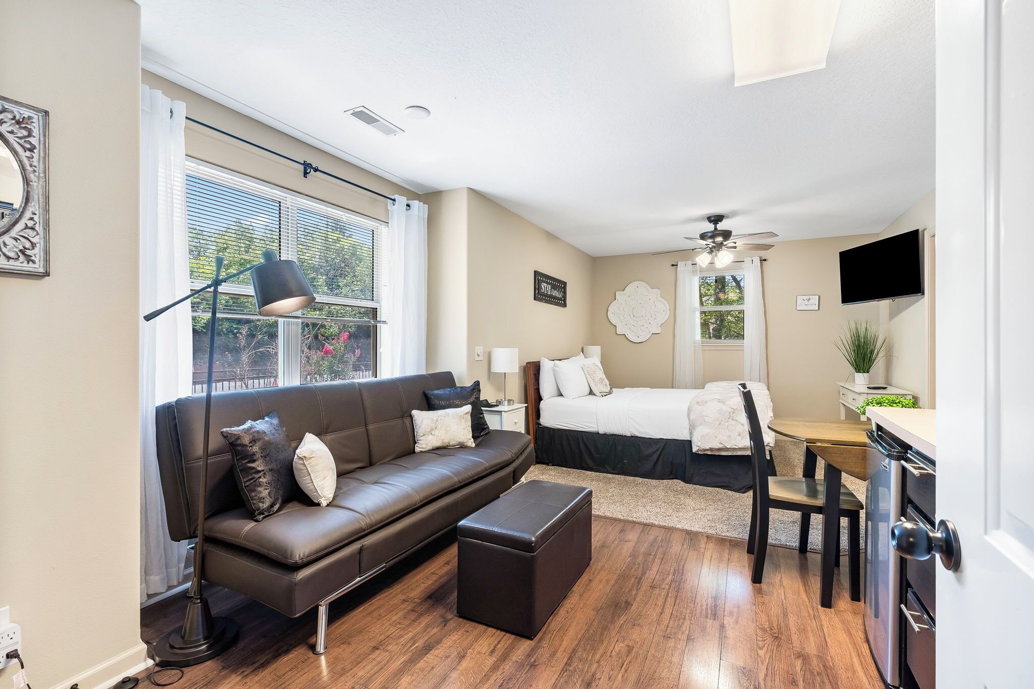 A modern, well-lit room with a brown leather sofa, a small ottoman, a dining table with chairs, a bed with a white headboard, and a ceiling fan.