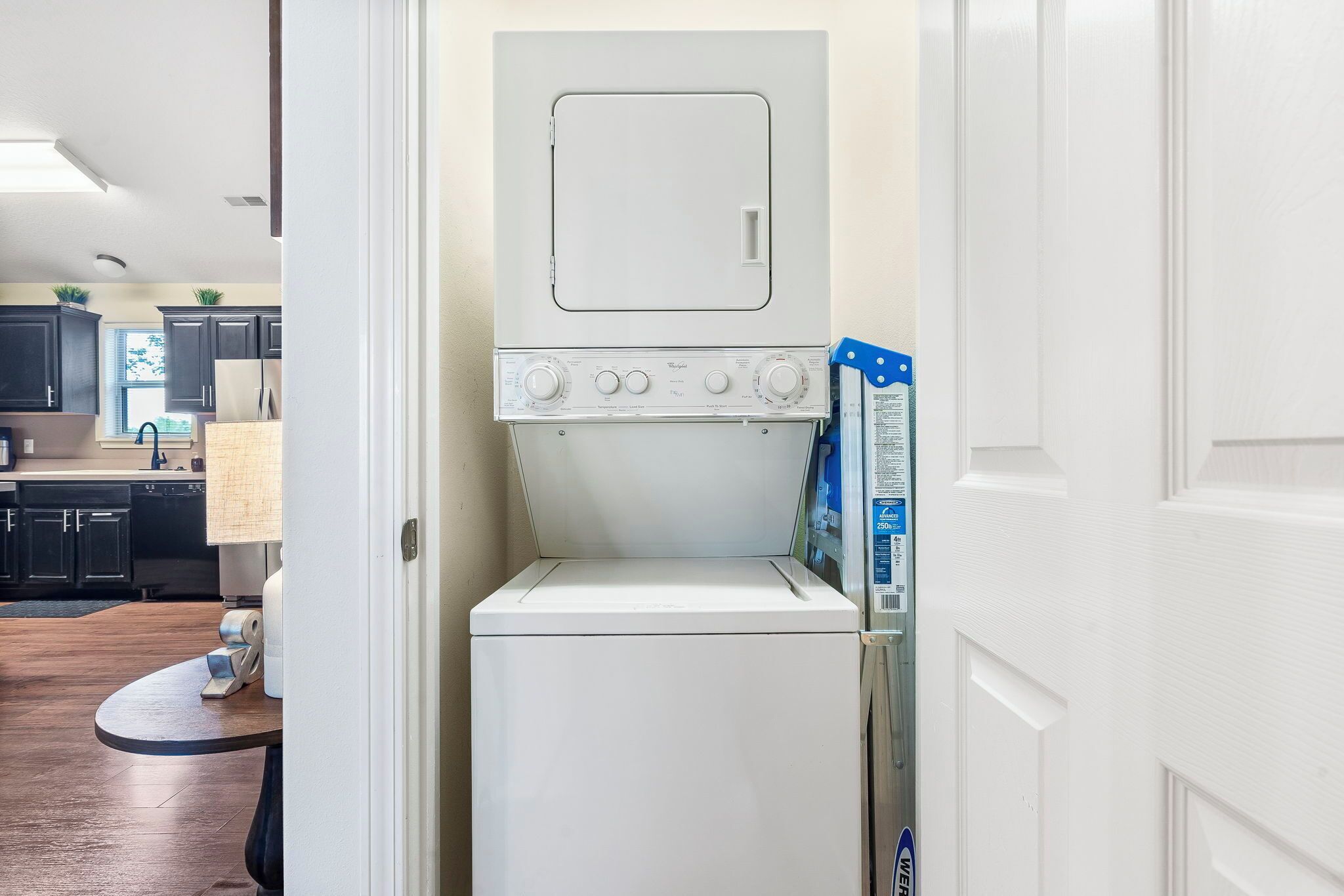 A compact laundry room with a front-loading washer and dryer, a small table with a towel, and a door leading to a kitchen area.