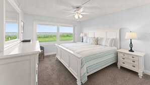 A bedroom with a bed, nightstands, a dresser, a ceiling fan, and large windows with a view of greenery. - Cozy Ground Floor Walkout w/Ocean View (Cocoa Beach)