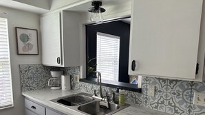 A modern kitchen with white cabinets, a marble countertop, a built-in sink, and a window with blinds. - Cozy 2/1 Close to Theme Parks! (St. Cloud)
