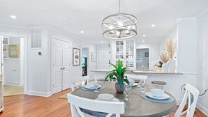 A dining area with a round table, white chairs, a chandelier, and a kitchen in the background.