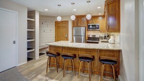 A modern kitchen with wooden cabinets, a stainless steel refrigerator, a microwave, and a coffee maker. There are four bar stools at the island.