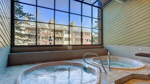A spa area with two hot tubs, a window with a view of buildings, and a tiled floor.
