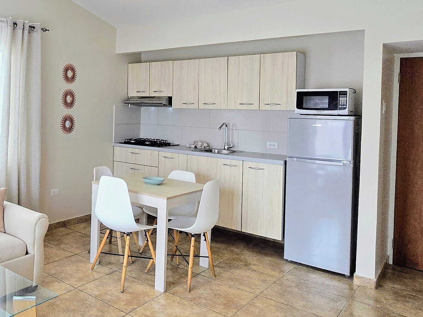 A compact kitchen with light wood cabinets, a stainless steel refrigerator, a microwave, and a dining area with white chairs and a glass table.
