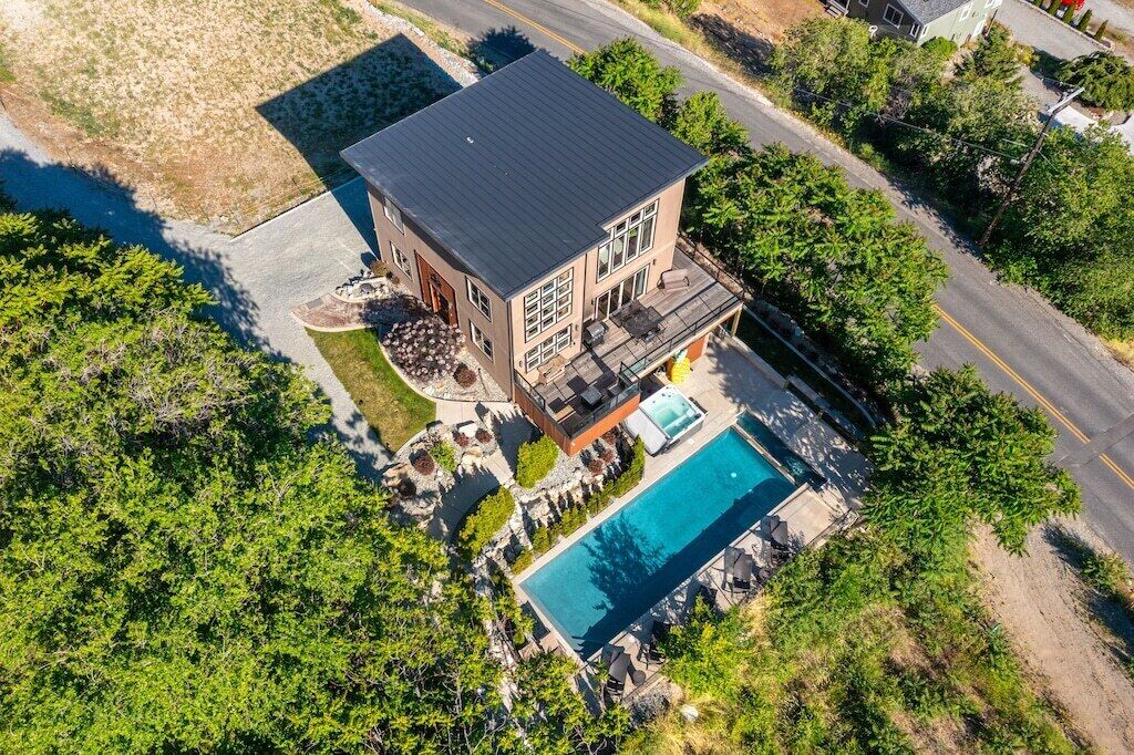 Vue aérienne d’une maison moderne à deux étages, dotée d’une piscine et entourée de verdure, avec une route à proximité.