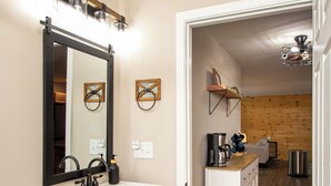 Un baño con tocador de madera oscura, un lavamanos blanco y un espejo. Hay un dispensador de papel higiénico y un cubo de basura. El baño conduce a una cocina con cafetera y estante.