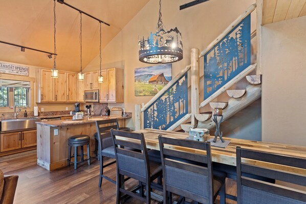 Une cuisine avec des meubles de rangement en bois, un îlot central doté de tabourets, un lustre et un escalier avec une rampe décorée sur le thème de la forêt.