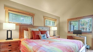 A bedroom with a large bed, two bedside tables with lamps, a desk with a computer, and windows with blinds.