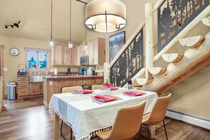 A dining area with a table set for two, wooden cabinets, a kitchen in the background, and a staircase with a unique design. - Private Hot Tub - Huge Covered Deck- Garage - On Ski Bus Route (Steamboat Springs)