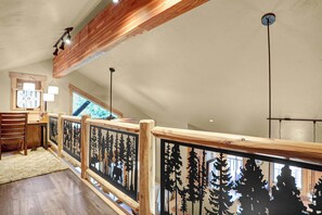 A wooden balcony with a railing featuring tree silhouettes, a desk with a chair, and a window with a view of greenery. - Private Hot Tub - Huge Covered Deck- Garage - On Ski Bus Route (Steamboat Springs)