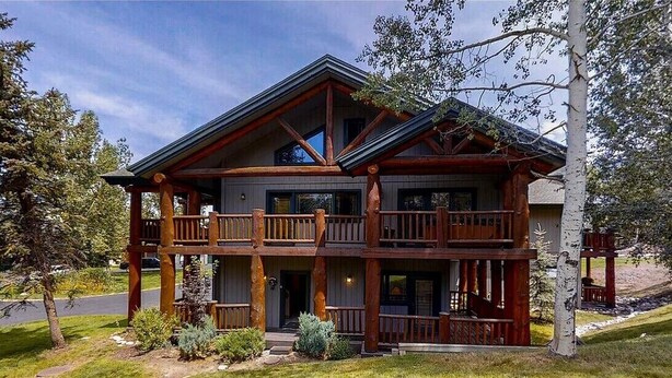 A two-story log cabin with a balcony on the upper floor, surrounded by trees and a well-maintained lawn.