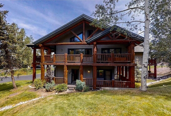 A two-story log cabin with a balcony on the upper floor, surrounded by trees and a well-maintained lawn. - Private Hot Tub - Huge Covered Deck- Garage - On Ski Bus Route (Steamboat Springs)