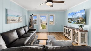 A living room with a black leather sofa, a wooden coffee table, and a TV mounted on the wall. There is a ceiling fan, a painting of a beach scene, and a view of a Ferris wheel through a door. - BEACHFRONT!!! Updated 2 Bedroom 2 Bath  Condo (Ocean Isle Beach)