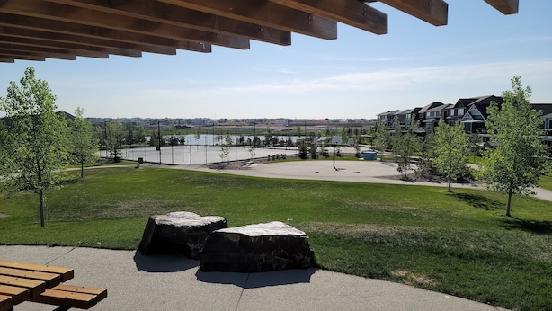 A park with a basketball court, picnic tables, and a view of a lake and residential buildings. - Luxurious home away from home with AC in a quiet & Clean Neighborhood just 4 U (Airdrie)