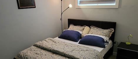Une chambre à coucher avec un lit, une lampe sur pied, une fenêtre avec des stores, un tableau au mur et un luminaire au plafond.