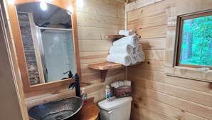 Une salle de bain rustique, avec des murs en bois, un grand miroir, des étagères en bois, des toilettes et une fenêtre donnant sur la forêt.
