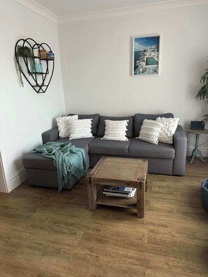 Sala de estar com sofá cinzento, almofadas brancas, mesa de centro de madeira e um quadro na parede.