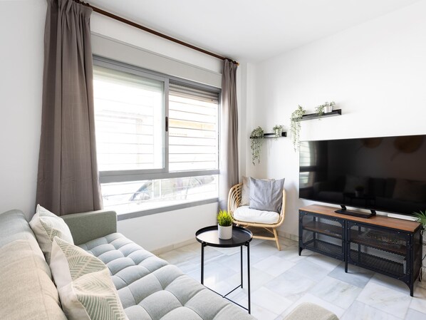 Una sala de estar moderna con un sofá gris, una mesa redonda de centro con una planta, un sillón de mimbre, un televisor sobre un mueble negro y una ventana con cortinas.