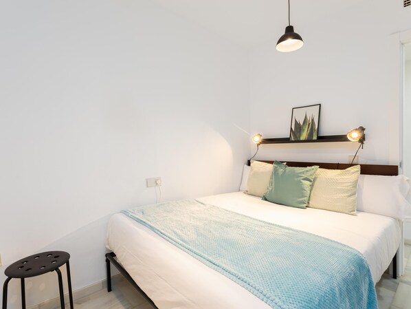 Un dormitorio ordenado con una cama, lámparas de noche, un taburete pequeño y un cuadro enmarcado en la pared.