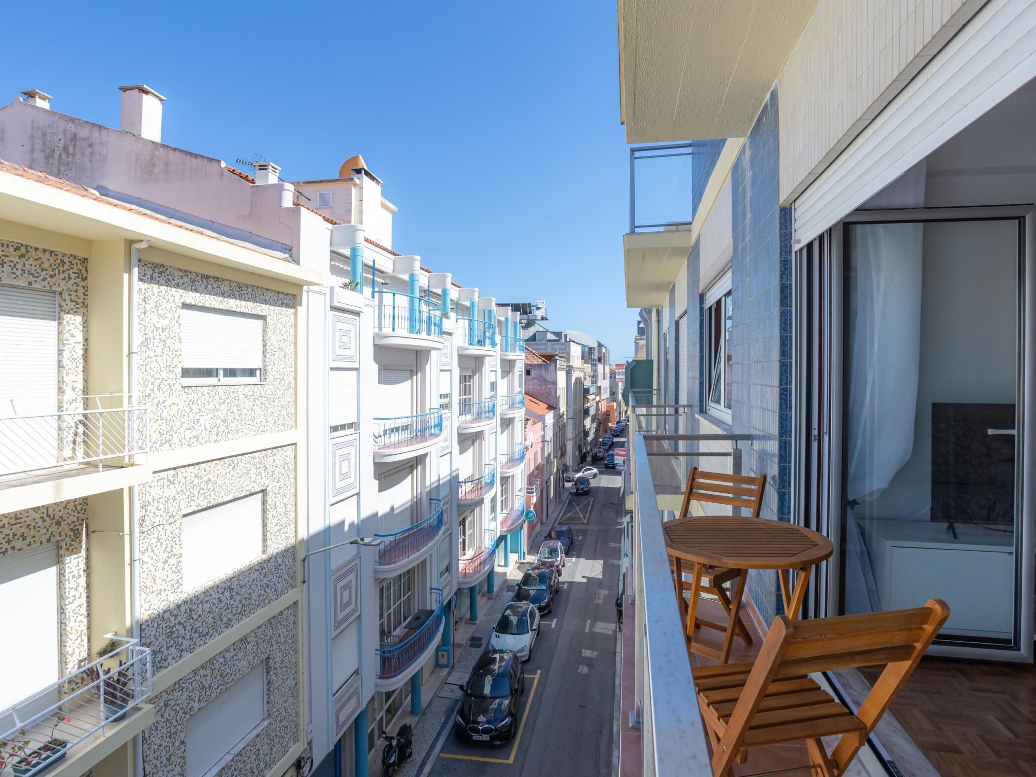 Un balcón con mesa y sillas de madera, con vista a una calle con coches aparcados y edificios con balcones.