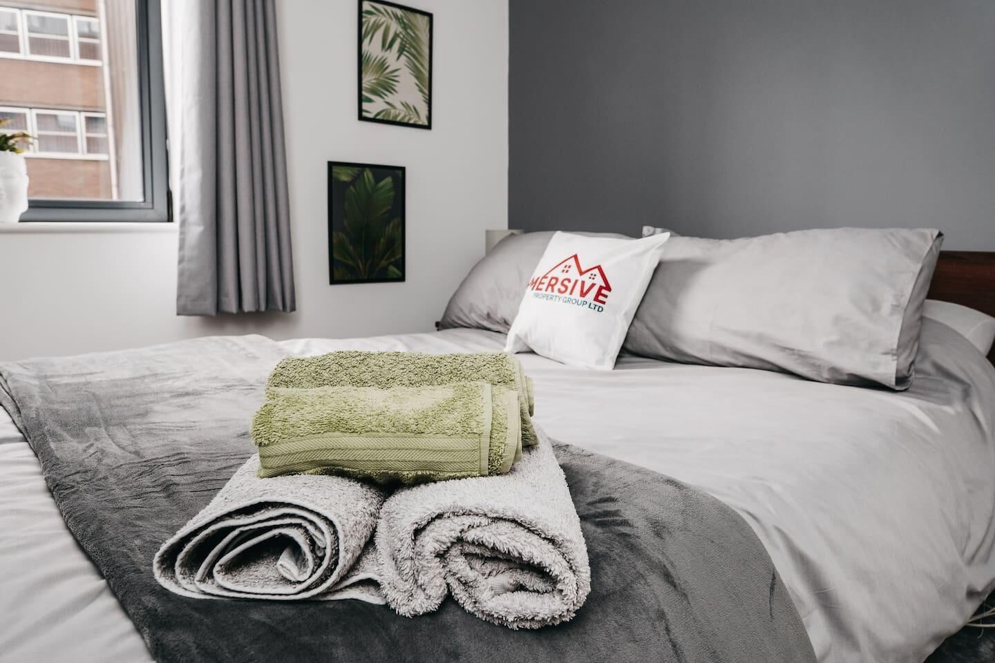Un lit bien fait, avec une serviette verte pliée et une serviette blanche roulée, une couverture grise, un oreiller orné d’un logo et des photos encadrées accrochées au mur.
