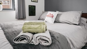 Un lit bien fait, avec une serviette verte pliée et une serviette blanche roulée, une couverture grise, un oreiller orné d’un logo et des photos encadrées accrochées au mur.