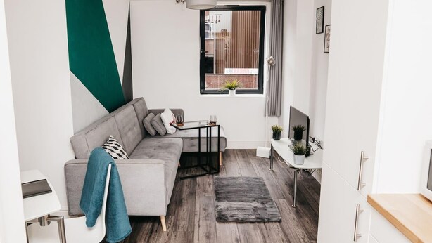 Un salon moderne avec un canapé gris, une petite table et un téléviseur. Il y a une fenêtre avec des rideaux et une vue sur l’extérieur.