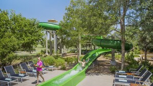 A water park with a green water slide, lounge chairs, and a person walking with a tray. - 1-Bedroom Suite at a 4⭐️ Hotel (San Antonio)
