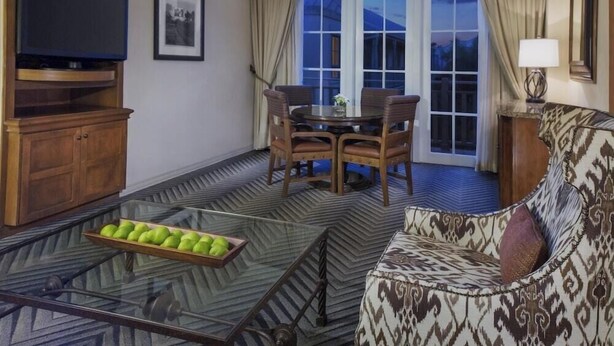 A living room with a glass coffee table, a sofa, a TV on a wooden stand, a dining table with chairs, and a large window with curtains. - 1-Bedroom Suite at a 4⭐️ Hotel (San Antonio)