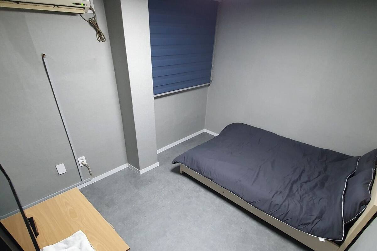 A small, sparsely furnished room with a single bed, a desk, and a window with blinds.
