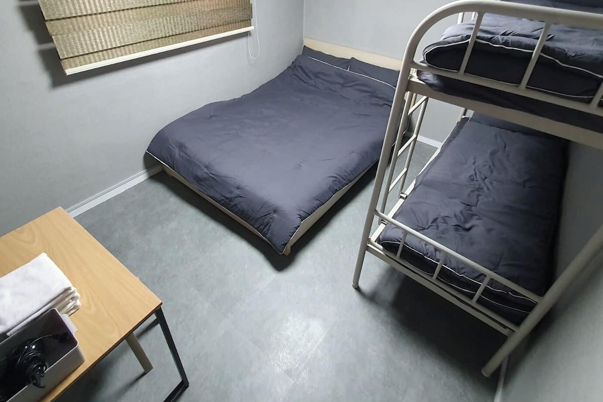 A dormitory room with bunk beds, a single bed, a table with a chair, a window with blinds, and an air conditioning unit.