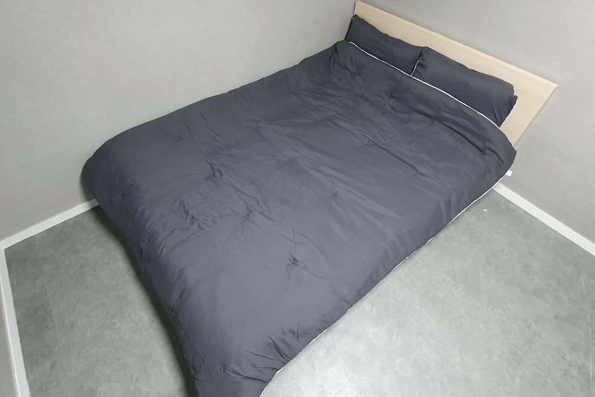 A bed with a grey duvet and wooden headboard in a room with grey walls and floor.