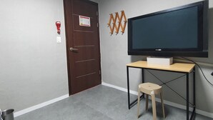 Um quarto com porta de madeira, televisão fixada na parede, um banquinho pequeno e um balde de metal no chão.
