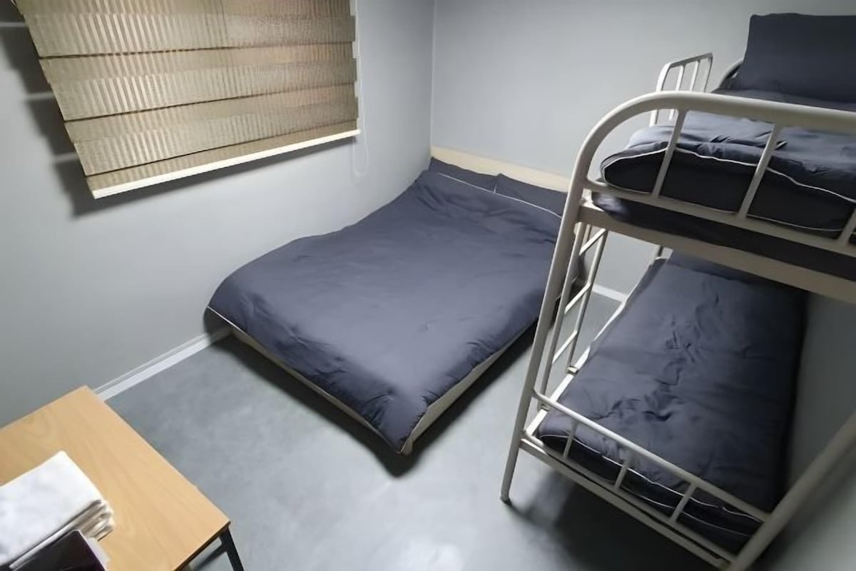 A single bed with a dark blue mattress and a metal bunk bed with matching bedding. A small wooden table with a white cloth is visible.