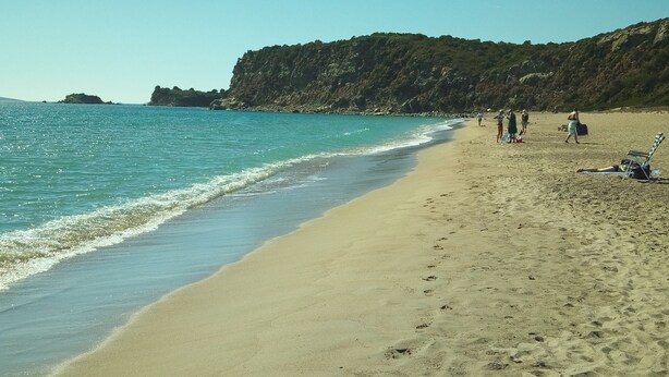 A beach with clear blue water, sandy shore, and rocky cliffs in the background.