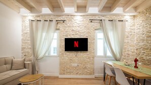 A cozy living room with a stone wall, a flat-screen TV, a sofa, a dining table with chairs, and a small wooden stool. - Casa di Marina in Corfu (Corfu)