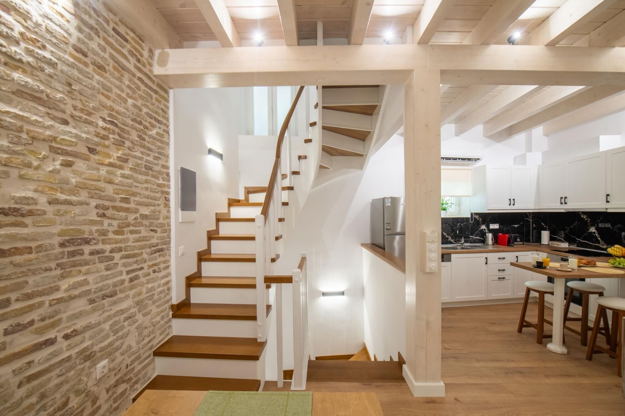 A modern kitchen with white cabinets, a wooden staircase, and a brick wall.