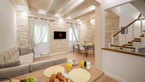 A cozy living room with a stone wall, wooden ceiling beams, a flat-screen TV, a dining area with a table set for two, and a staircase. - Casa di Marina in Corfu (Corfu)