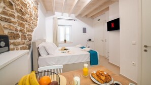 A small, cozy room with a stone wall, a bed, a TV, and a breakfast table with fruits and drinks. - Casa di Mana in Corfu (Corfu)