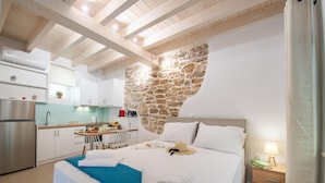 A bedroom with a stone wall, a bed with white linens, a bedside table with a lamp, and a view of the kitchen with white cabinets and a refrigerator. - Casa di Mana in Corfu (Corfu)