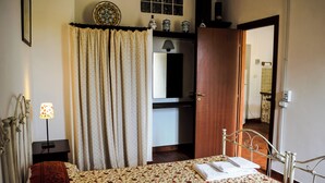 Um quarto com cama, mesa de cabeceira, luminária, janela com cortinas, espelho e porta para outro cômodo.